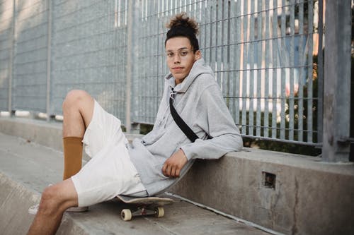 年轻的种族小伙子坐在城市公园篱笆附近的滑板 · 免费素材图片