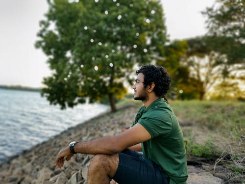坐在水附近的岩石上的绿色t恤和蓝色牛仔牛仔裤的男人 · 免费素材图片