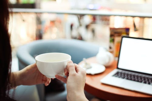 在macbook Pro面前拿着白色陶瓷茶杯的人 · 免费素材图片