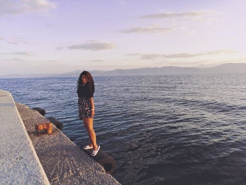 黑色上衣和花卉短裤站在海边的岩石上的女人 · 免费素材图片