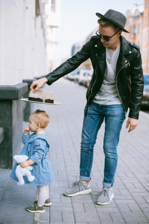 黑色皮夹克的男人抱着婴儿的蓝色礼服站在人行道上 · 免费素材图片