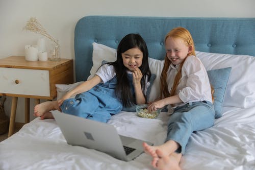 性格开朗的小多民族女友在笔记本电脑上看电影，在床上吃爆米花 · 免费素材图片