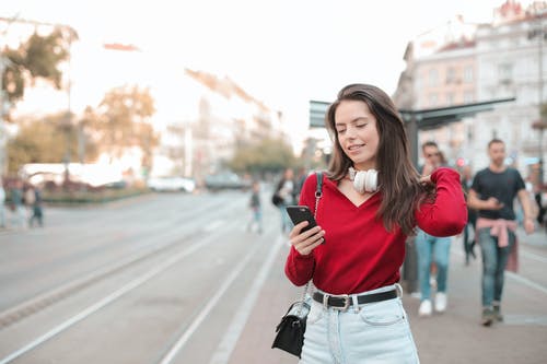 女人穿着红色毛衣和蓝色牛仔牛仔裤，站在人行道上时使用她的手机的选择性聚焦照片 · 免费素材图片