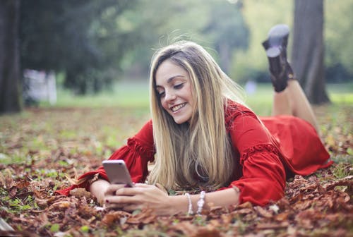 在发短信时躺在干树叶的肚子上的红色长袖衬衫的微笑妇女的照片 · 免费素材图片