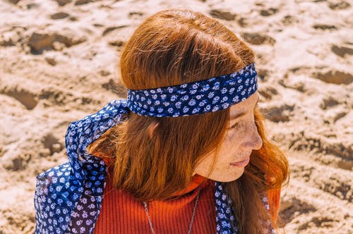 坐在蓝色的花头巾和橙色的顶部，坐在沙滩上看着别处的雀斑的女人的照片 · 免费素材图片