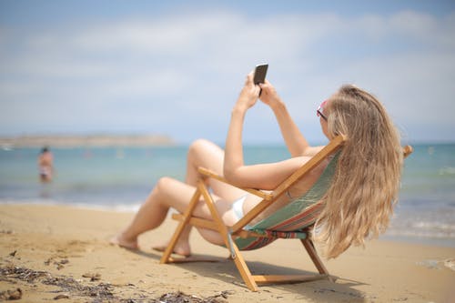 蓝色和白色比基尼的女人坐在沙滩上的棕色木椅上，同时使用智能手机 · 免费素材图片
