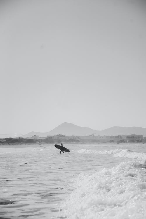 携带冲浪板在水上行走的人的灰度照片 · 免费素材图片