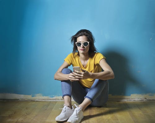 女人穿着黄色t恤，蓝色牛仔裤和白色太阳镜坐在蓝色墙前的木地板上的照片 · 免费素材图片