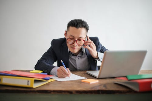 蓝色西装外套，条纹的衬衫和眼镜坐在一张桌子和他的笔记本电脑在电话上交谈的人的照片 · 免费素材图片