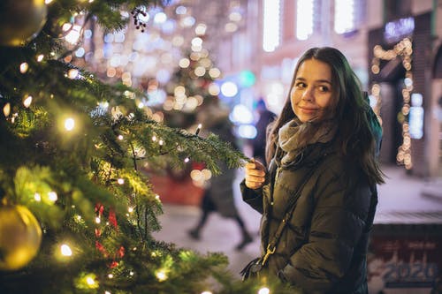 泡泡夹克站在圣诞树与弦灯附近的女人 · 免费素材图片