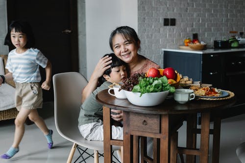 积极的小亚洲女孩跑在奶奶和哥哥后面在餐桌旁拥抱在早餐期间 · 免费素材图片