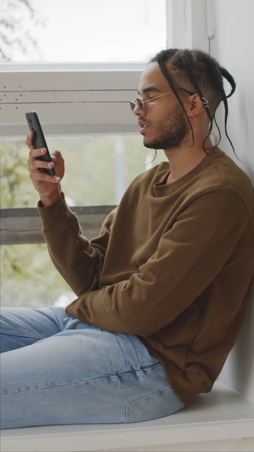 一个年轻人坐在窗台上时使用他的电话 · 免费素材视频